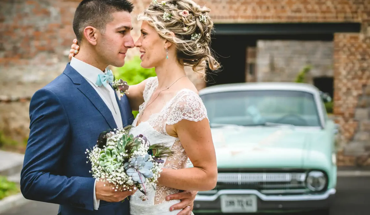 couple de mariés posant pour un shooting et dirigé par un photographe. Ils sont enlacés, tiennent un bouquet de fleurs entre eux et se regardent amoureusement dans les yeux. En arrière pla, on devine une voiture verte pale de style années 60