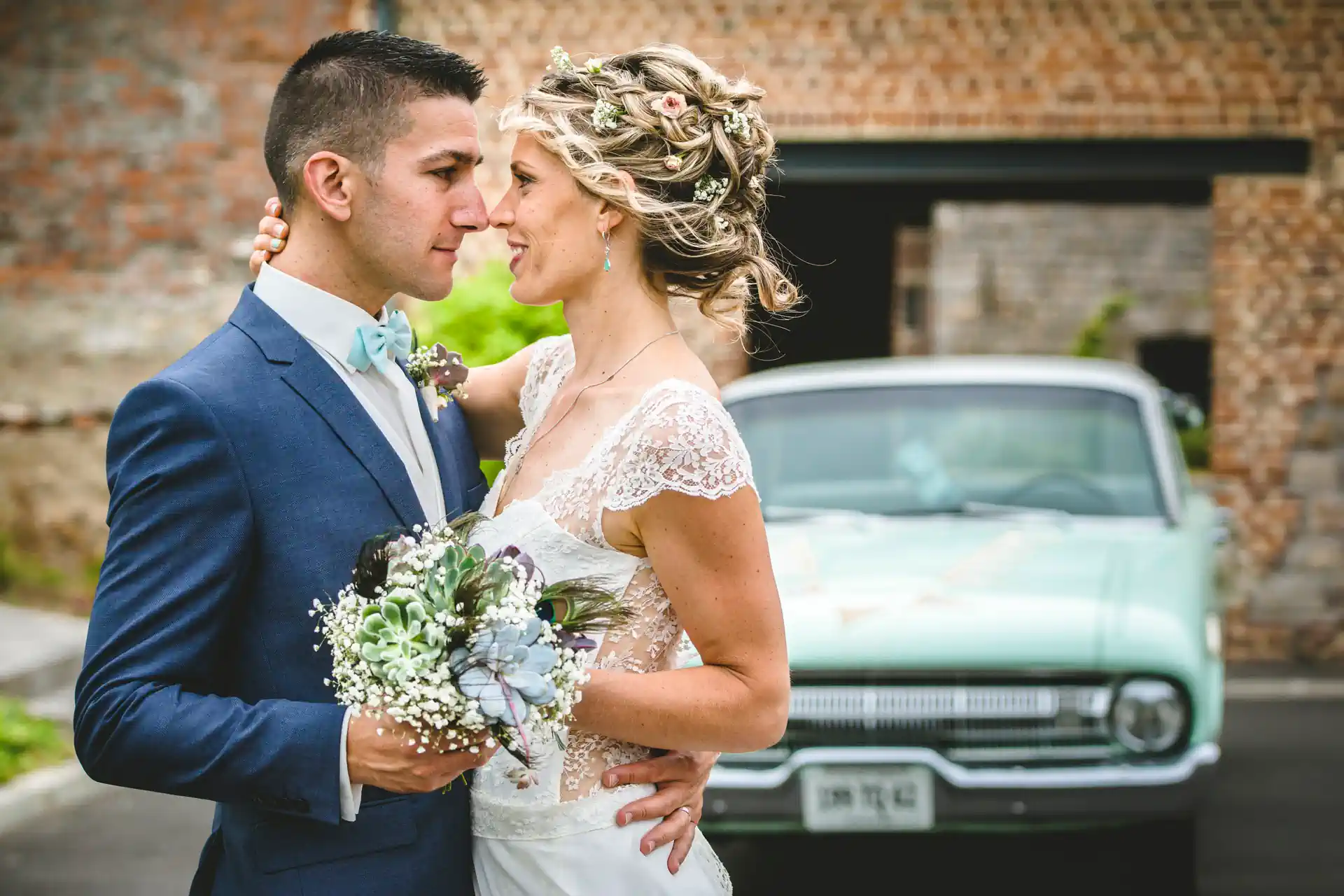 couple de mariés posant pour un shooting et dirigé par un photographe. Ils sont enlacés, tiennent un bouquet de fleurs entre eux et se regardent amoureusement dans les yeux. En arrière pla, on devine une voiture verte pale de style années 60