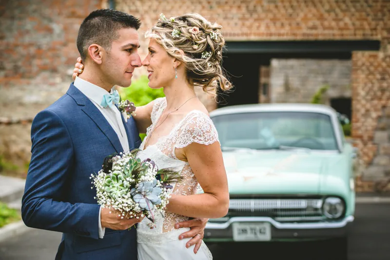 couple de mariés posant pour un shooting. Ils sont enlacés, tiennent un bouquet de fleurs entre eux et se regardent amoureusement dans les yeux. En arrière pla, on devine une voiture verte pale de style années 60