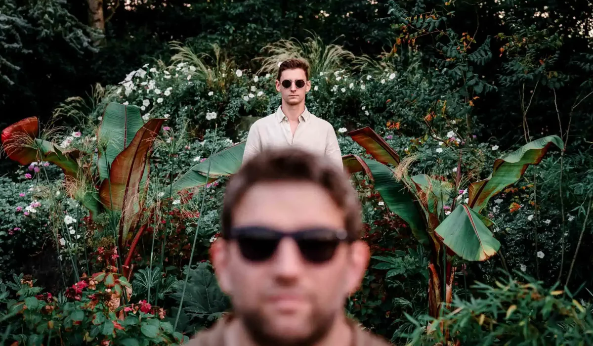 couple d'hommes avec des lunettes de soleil posant dans un parc entourés d'une plantes et de fleurs