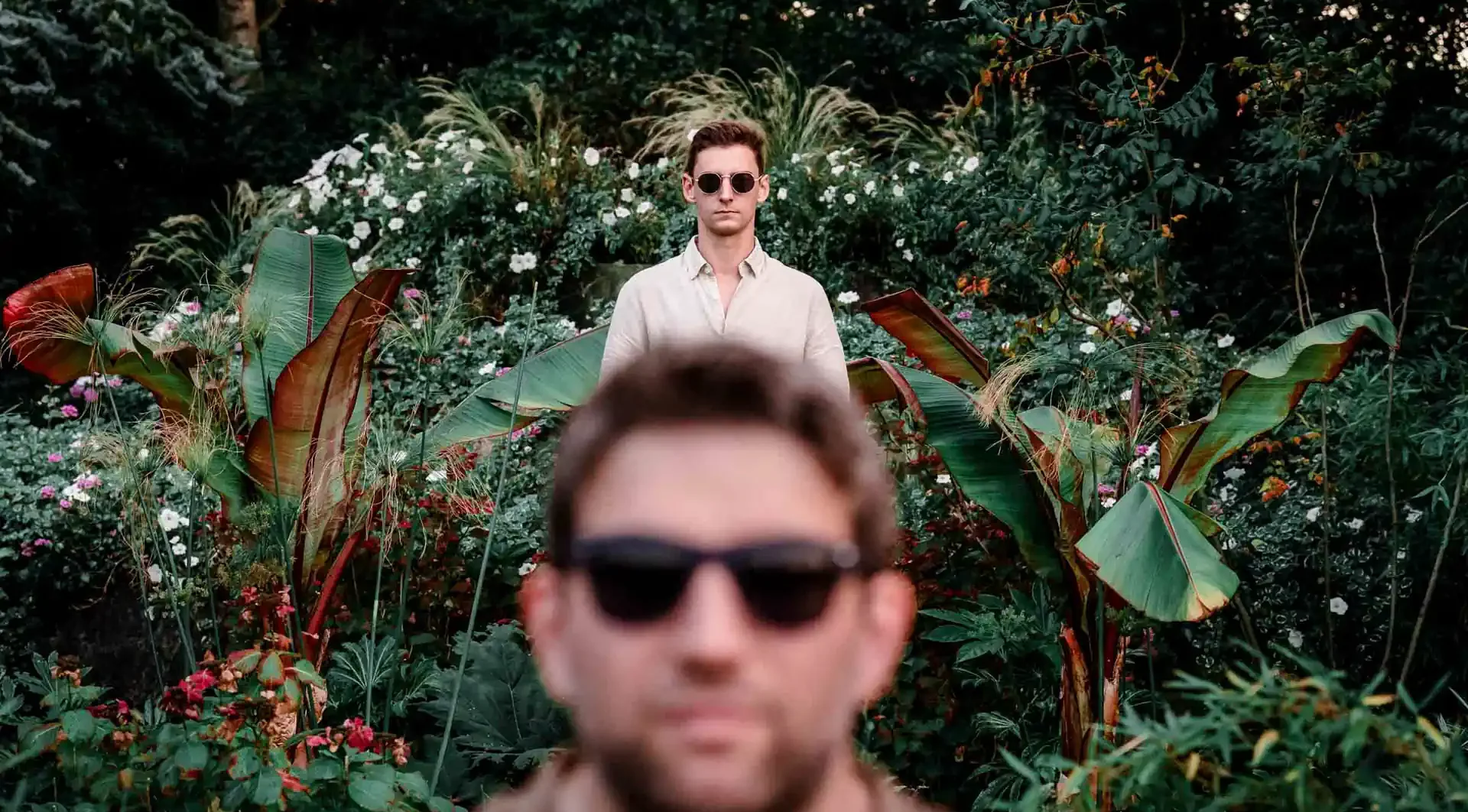 couple d'hommes avec des lunettes de soleil posant dans un parc entourés d'une plantes et de fleurs