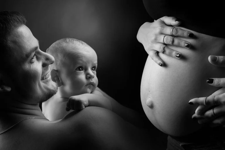 photo noir et blanc prise en studio d'un père qui tient son bébé dans les bras près du ventre d'une femme enceinte