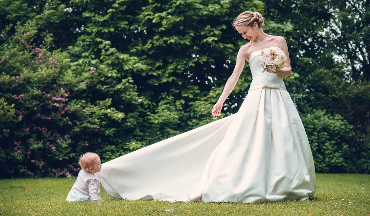 un bébé assis dans l'herbe tire la longue robe d'une mariée qui lui sourit