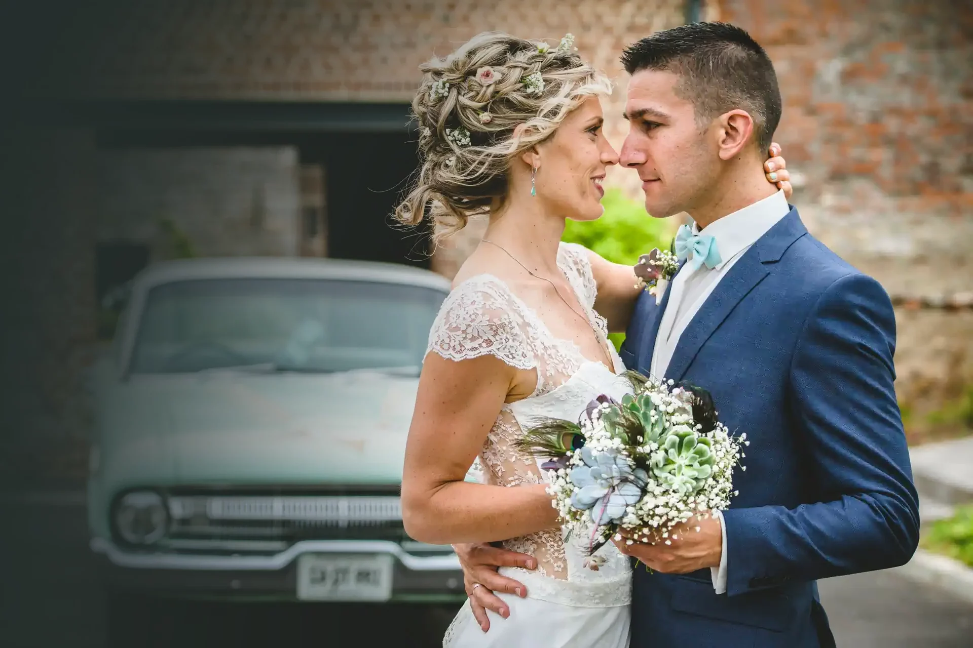 couple de mariés posant pour un shooting et dirigé par un photographe. Ils sont enlacés, tiennent un bouquet de fleurs entre eux et se regardent amoureusement dans les yeux. En arrière pla, on devine une voiture verte pale de style années 60