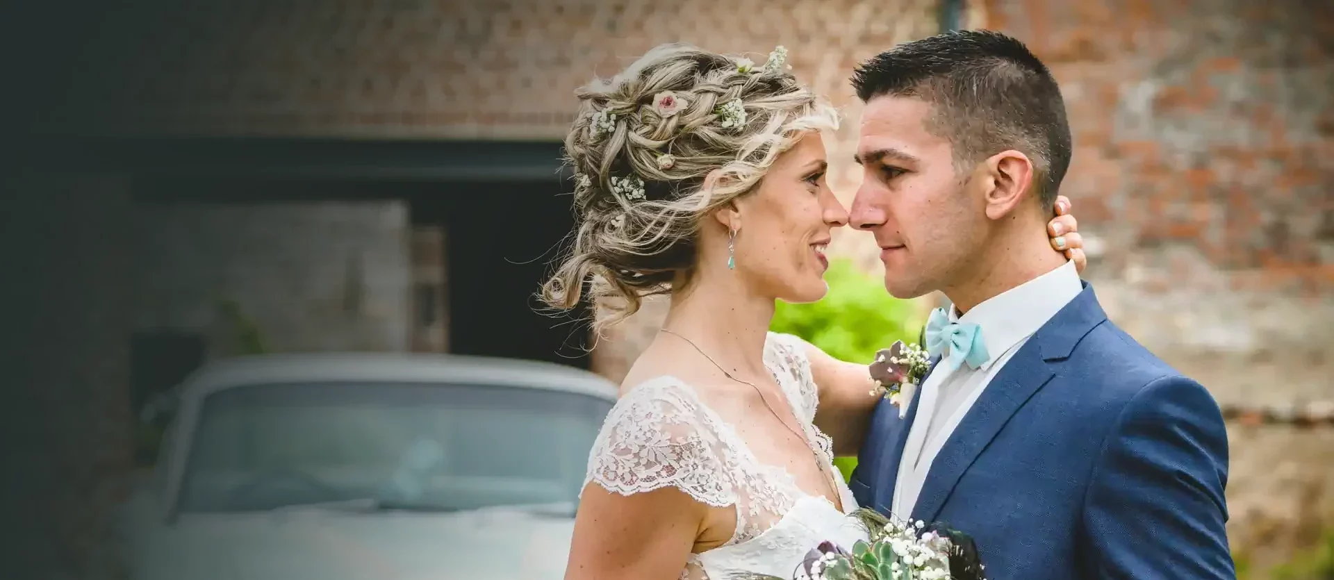 couple de mariés posant pour un shooting et dirigé par un photographe. Ils sont enlacés, tiennent un bouquet de fleurs entre eux et se regardent amoureusement dans les yeux. En arrière pla, on devine une voiture verte pale de style années 60