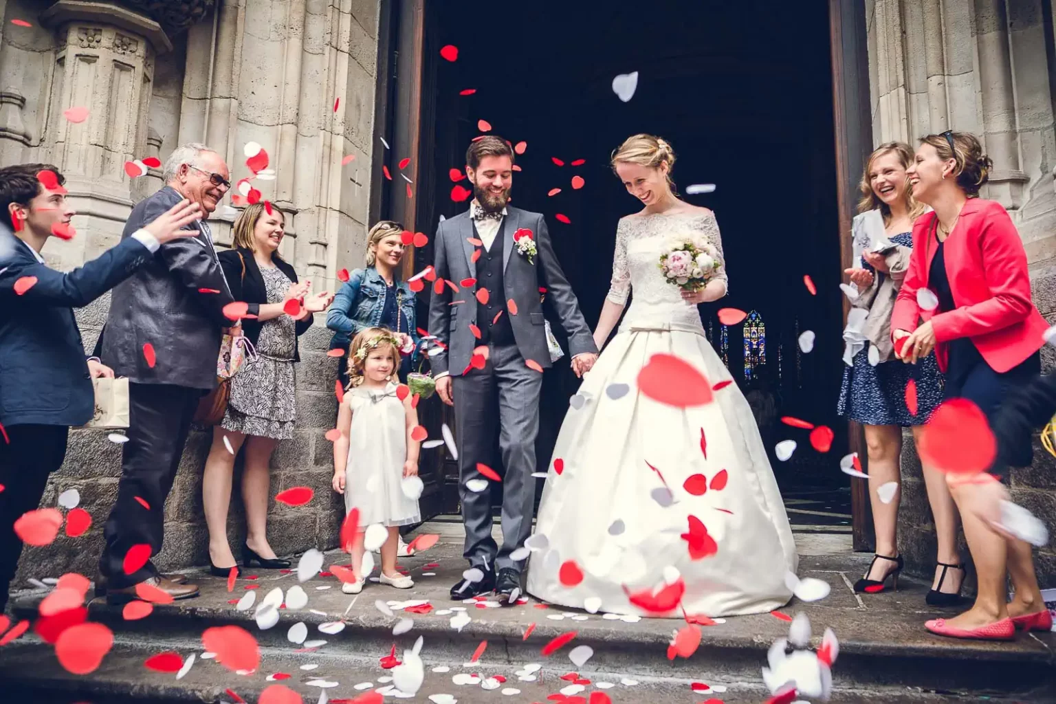 photo mariage d'un couple sortant de l'église entouré de leurs familles qui leur jettent des pétales de roses rouges.