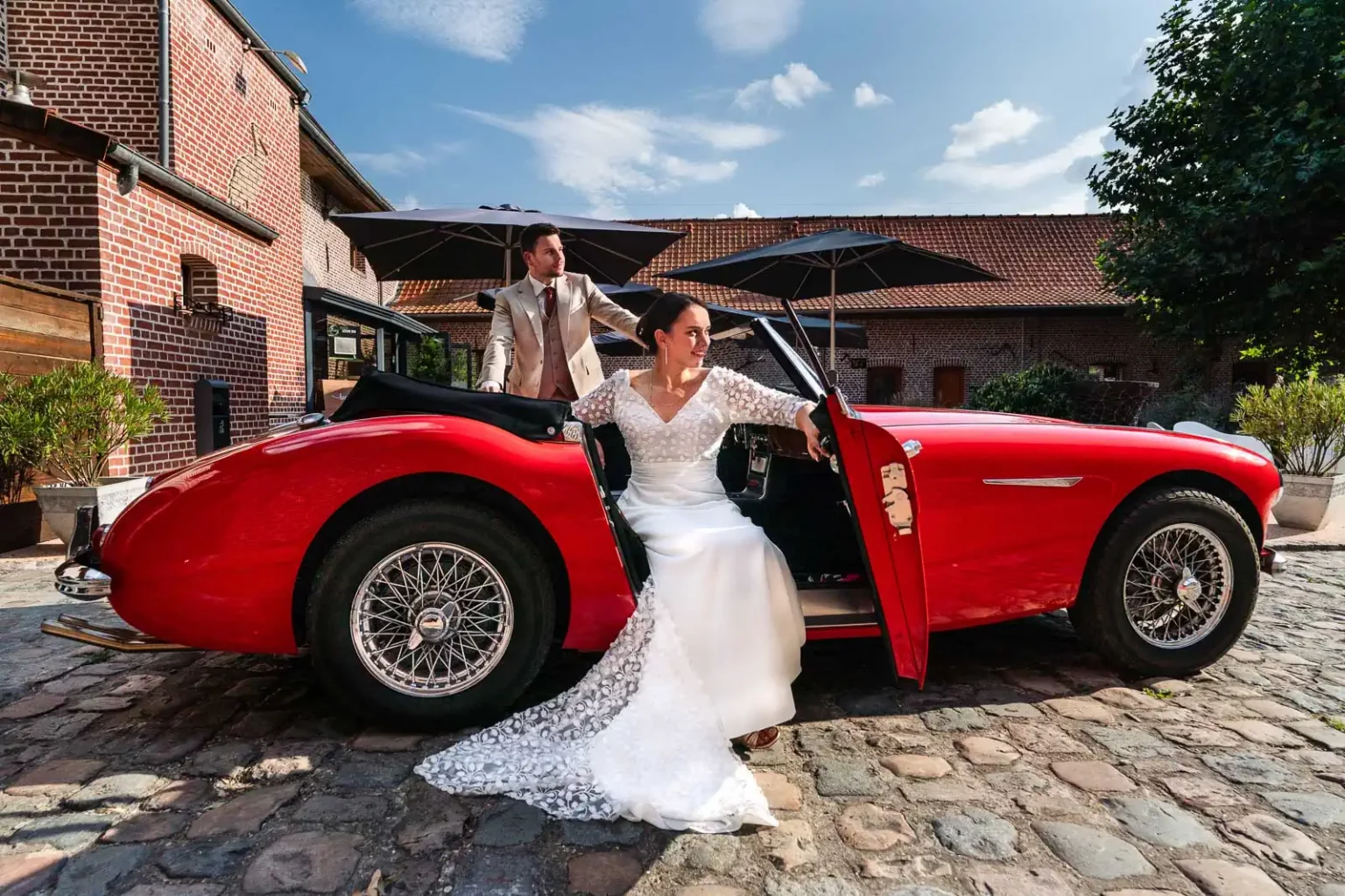 photo de mariage d'un couple posant sur une voiture rouge dans la cour intérieure du Domaine des Cigognes