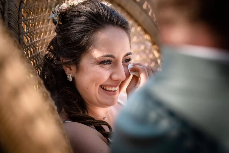 photo de la mariée qui pleure d'émotion lors de la cérémonie laïque. Elle sèche ses larmes avec un mouchoir tout en souriant. Elle est assise dans un grand fauteuil en osier