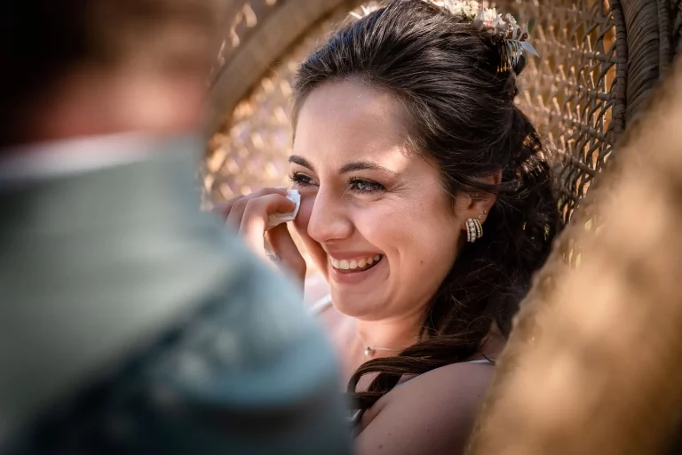 photo de la mariée qui pleure d'émotion lors de la cérémonie laïque. Elle sèche ses larmes avec un mouchoir tout en souriant. Elle est assise dans un grand fauteuil en osier