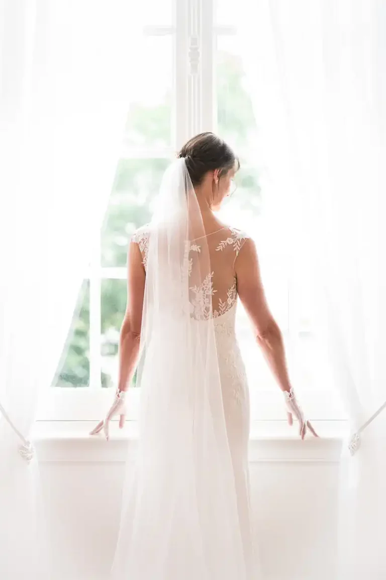 photo d'une femme en robe de mariée vue de dos. Elle pose devant une fenêtre dans une atmosphère lumineuse