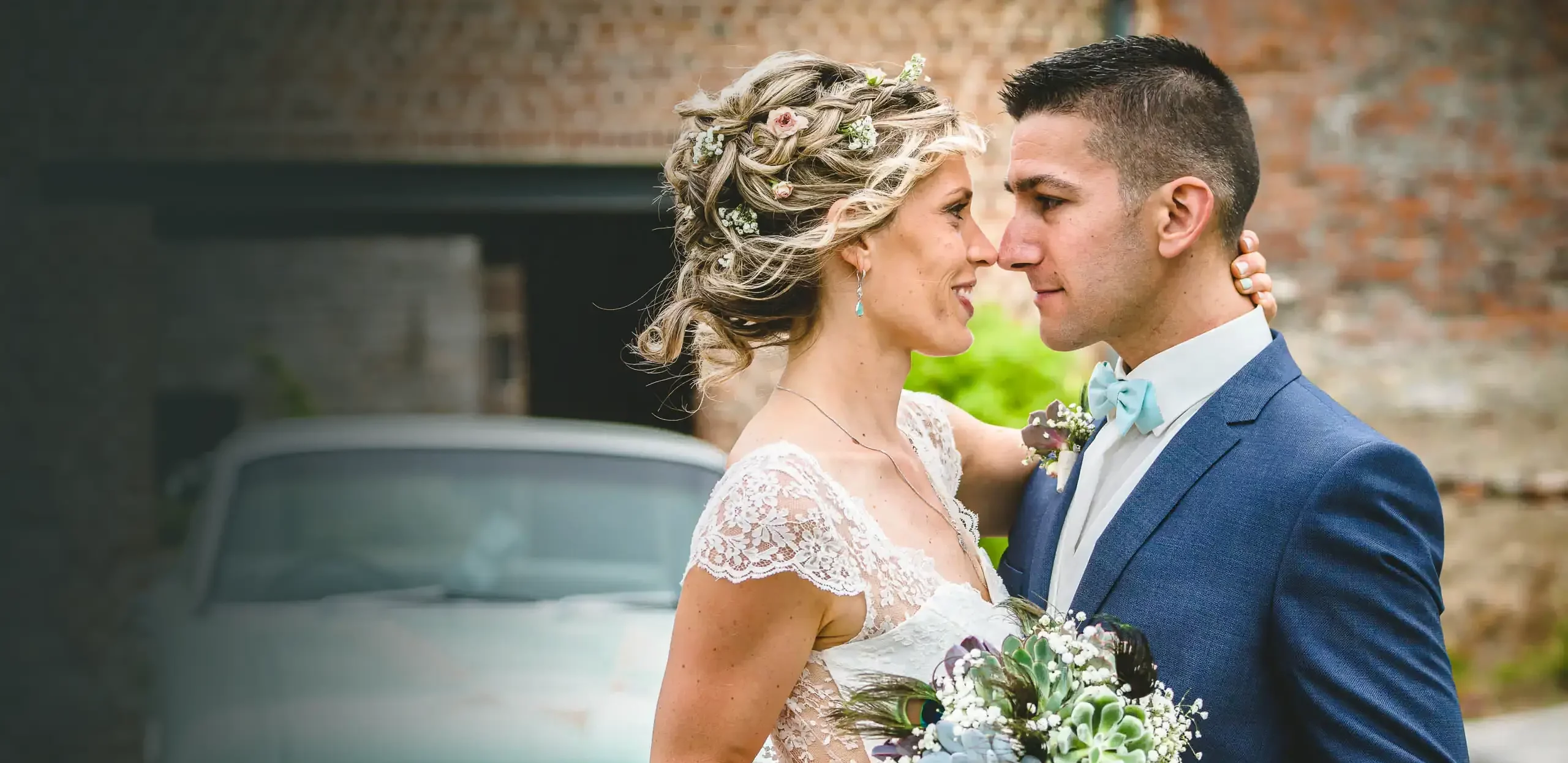 couple de mariés lors d'un shooting photo dirigé par un photographe. Ils sont enlacés, tiennent un bouquet de fleurs entre eux. En arrière plan, une voiture verte de style années 60