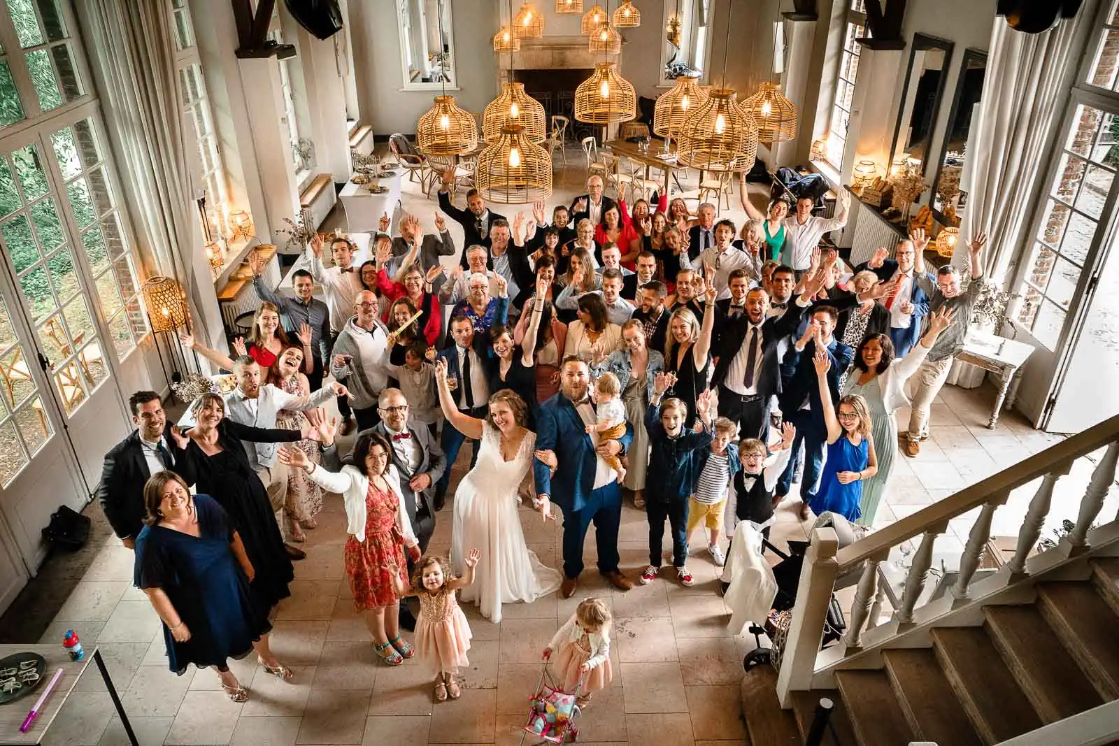 photo de groupe lors d'un mariage avec les mariés au centre entouré de tous leurs invités. La photo est prise en plongée à partir du premier étage, donnant une vue large sur toute la salle de réception du domaine d'Achelles.